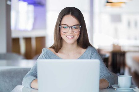 Portrait of cheerful Caucasian female student wearing eyeglasses using laptop in cafe for studies or networking and smiling